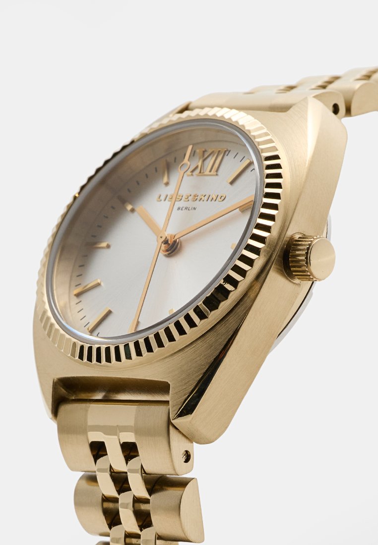 Goudkleurig horloge met een gestructureerde bezel, zilveren wijzerplaat en Romeinse cijfers. Kenmerkt zich door een schakelband en minimalistische uurmarkeringen.