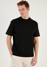 Tricou din bumbac negru, cu guler rotund și mâneci scurte, având o croială lejeră și o textură fină.