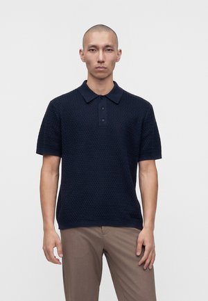 Uomo con testa rasata che indossa una polo blu navy a maniche corte con trama e pantaloni marrone chiaro su uno sfondo chiaro uniforme.