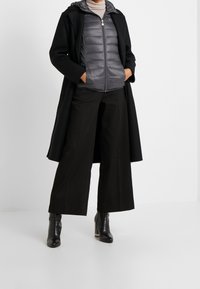 Abrigo largo negro sobre una chaqueta acolchada gris, combinado con pantalones anchos negros y botas negras hasta el tobillo. Superficies texturizadas y capas bien definidas.