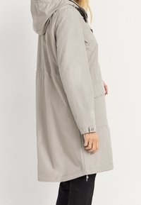 Vaaleanharmaa vedenpitävä takki, jossa on huppu, säädettävä vyötärö, sivutaskut ja napilliset hihansuut; sileä, tekstuuri pintakäsittely.