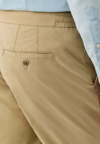 Detailný záber na béžové nohavice s zapnutým zadným vreckom, nosené s jemne modrou košeľou zastrčenou dovnútra.