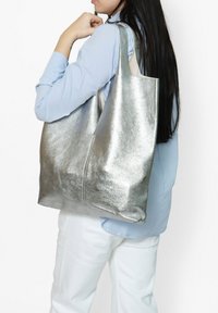 Metalen zilveren draagtas met een gladde textuur, groot open ontwerp en zachte handvatten. Casual gedragen over een lichtblauw shirt.
