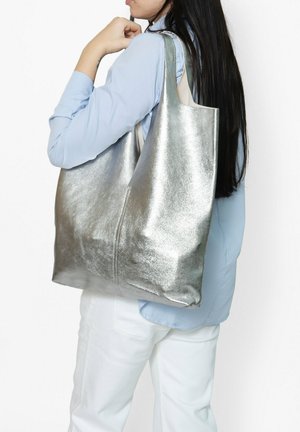 Borsa tote argento metallico con una texture liscia, design ampio e manici morbidi. Indossata in modo casual sopra una camicia azzurro chiaro.