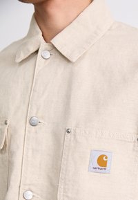 Beige canvas jacket with a pointed collar, silver snap buttons, two chest pockets, and a blue and orange Carhartt logo patch.
