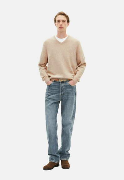 Suéter de lana beige claro con escote en V sobre una camiseta blanca, combinado con jeans azul de pierna recta y zapatos de ante marrones, con las manos en los bolsillos.