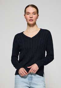 Marinblå kabelstickad tröja med V-ringning, långa ärmar och ribbade manschetter. Har en diskret logotyp på bröstet och en något förlängd bakdel.