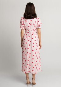 Kobieta z włosami do ramion w kolorze brązowym, ubrana w różową sukienkę midi z czerwonym nadrukiem kwiatowym i bufiaste rękawy, stojąca tyłem, widoczne beżowe szpilki.