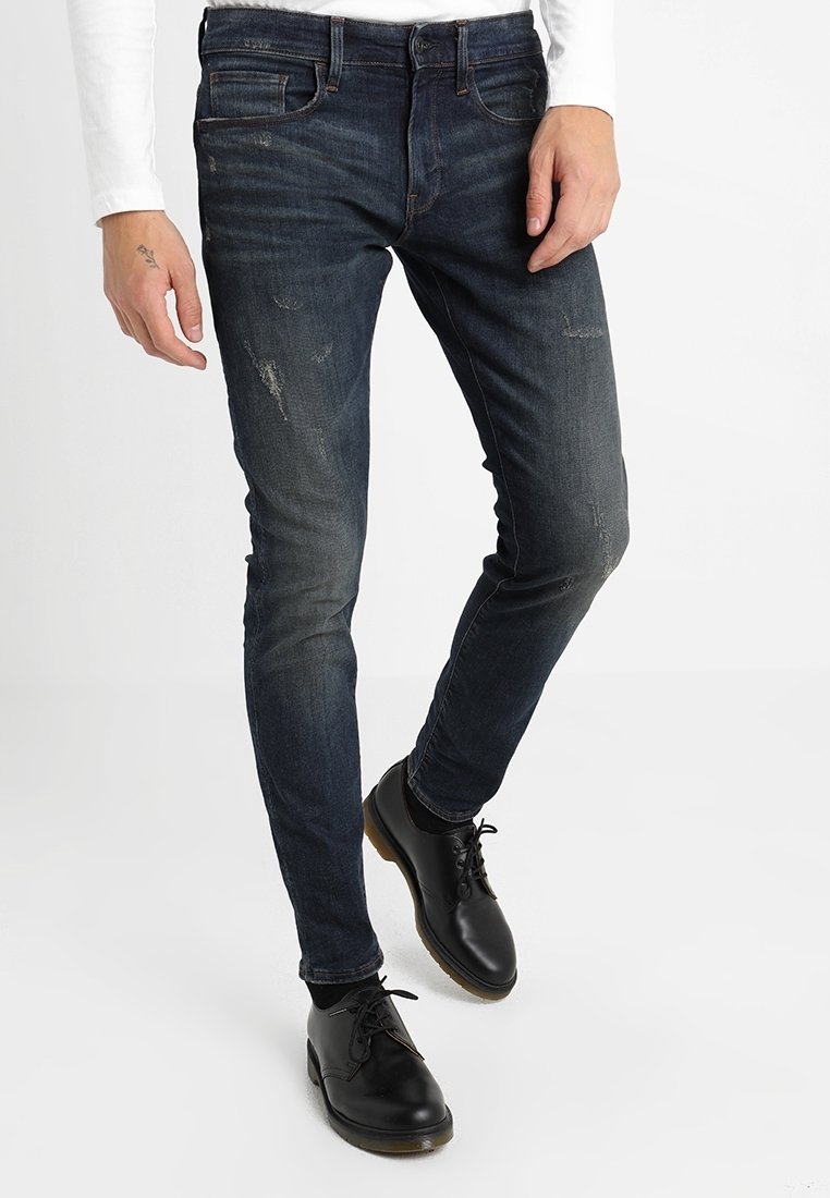 Mörkblå denimjeans med slim fit, med slitna detaljer och femficksdesign. Bärs tillsammans med svarta snörskor och en vit långärmad skjorta.