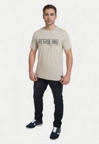 Lys beige t-shirt med teksten "PETROL IND" i mørk skrift. Bæres med sorte bukser og marineblå sneakers med hvide snørebånd.