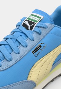 Tênis de camurça azul claro com detalhes em couro amarelo, atacadores texturizados e uma etiqueta da marca em preto e verde. Apresenta texto do logótipo em dourado.