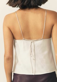 White satin camisole with thin straps, featuring a tie-back design. Smooth texture and loose fit, with a straight hemline at the bottom.