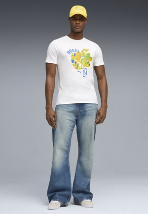 Man draagt een wit T-shirt met een Braziliaanse print, verbleekte wijde jeans, witte sneakers en een gele pet tegen een grijs verloopachtergrond.