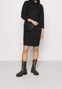 Czarna sukienka z dekoltem typu cowl i długimi rękawami, zestawiona z czarną okrągłą torebką na ramię oraz oliwkowymi botkami za kostkę o teksturowanej podeszwie.