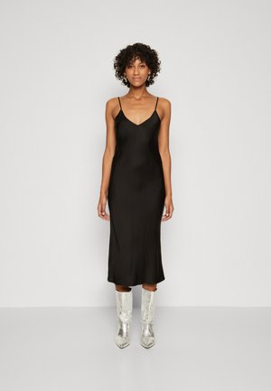 Abito slip in raso nero con spalline sottili, lunghezza midi e una texture liscia; abbinato a stivali al ginocchio in argento lucido.
