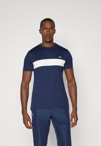 Maglietta blu navy a maniche corte, con una striscia orizzontale bianca e un accento verde. Collo rotondo con un piccolo logo sul petto.