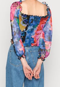Blusenshirt mit floralem Muster, transparenten, mehrfarbigen Ärmeln und gerüschtem Oberteil. Rüschenmanschetten mit schwarzem Rand. Kombiniert mit hochgeschnittenem Jeans.