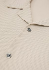 Camisa de tela texturizada beige con cuello vuelto y dos botones redondos plateados visibles en la parte delantera.