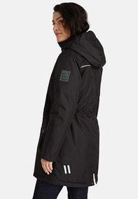 Vrouw in een zwarte gewatteerde parka met een "URBAN WEAR" patch op de mouw en reflecterende strepen nabij de zoom, met haar gezicht naar links.