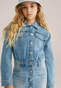 Gaišmataina meitene, ģērbusies gaiši zilos džinsu jaceļos ar priekšējiem pogām un gaiši zilu cepurīti, pret bēša fona.