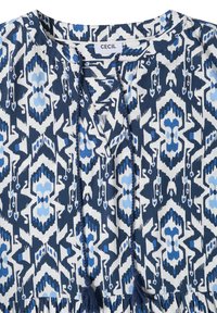 Blouse à motif géométrique bleu et blanc avec encolure ronde, ouverture devant et liens tressés à pompons, étiquette "CECIL" au col.