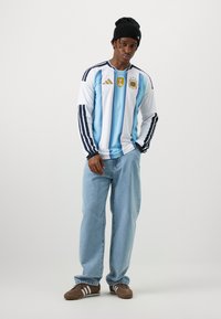 Ung man som bär Argentinas fotbollströja, ljusblå jeans, bruna Adidas-sneakers, svart mössa, stående mot vit bakgrund.
