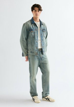 Ung mand iført lys blå denimjakke og jeans med hvid T-shirt samt hvide sneakers med sorte striber, stående mod en ensfarvet baggrund.