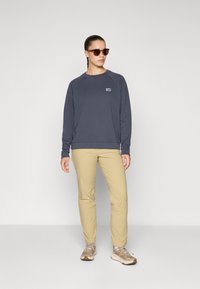 Mulher vestindo um moletom cinza escuro, calças bege, tênis bege e óculos de sol, em pé contra um fundo cinza claro liso.