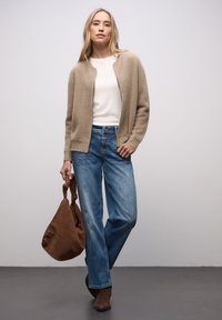 Cardigan beige en tricot porté sur un haut blanc, jean bleu ample, bottines en daim marron et un sac marron à texture douce.