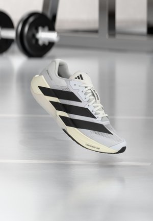 Chaussure de course Adidas blanche et noire avec trois bandes noires et semelle "LIGHTSTRIKE PRO", près d'un haltère de gym flou sur un sol brillant.