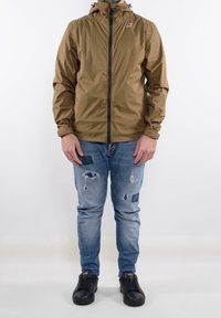 Brązowa, lekka kurtka z kapturem, przednim zamkiem błyskawicznym i logo. Noszona na podniszczonych niebieskich dżinsach i czarnych trampkach. Gładka tekstura, sportowy design.