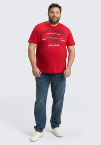 Rode katoenen T-shirt met zwarte tekst en grafisch ontwerp, gedragen met blauwe denim jeans en witte sneakers. Casual pasvorm, korte mouwen en ronde hals.