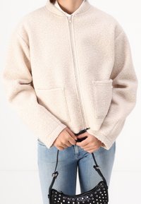 Beige fleecejacka med hög krage, framdragkedja och två fickor med lappar. Modellen håller en liten svart väska med silvernitar.