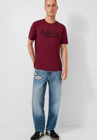 Burgundijska pamučna majica s crnim logom na prsima, uparena s svjetloplavim širokim trapericama s izrezom i crnim tenisicama.