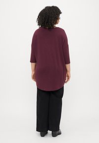 Top burgundy de corte solto com mangas de três quartos, bainha arredondada e combinado com calças largas pretas. Textura de tecido suave, design minimalista.