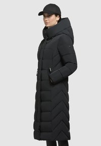 Giacca cushy nera con cappuccio, caratterizzata da un motivo a zigzag, tasche con zip e colletto alto, indossata con un berretto nero.