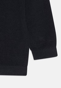 Maglione blu scuro con un motivo a coste testurizzato, con polsini e orlo aderente. Il tessuto appare spesso e caldo, adatto per essere indossato a strati.