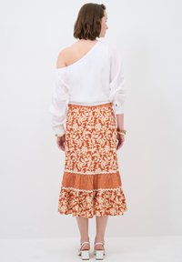 Blouse blanche à épaules dénudées avec des manches longues, associée à une jupe orange à motifs avec des designs floraux et un ourlet volanté.