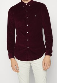 Hombre vistiendo una camisa de pana roja oscura con botones blancos y pantalones de colores claros, de pie contra un fondo liso.