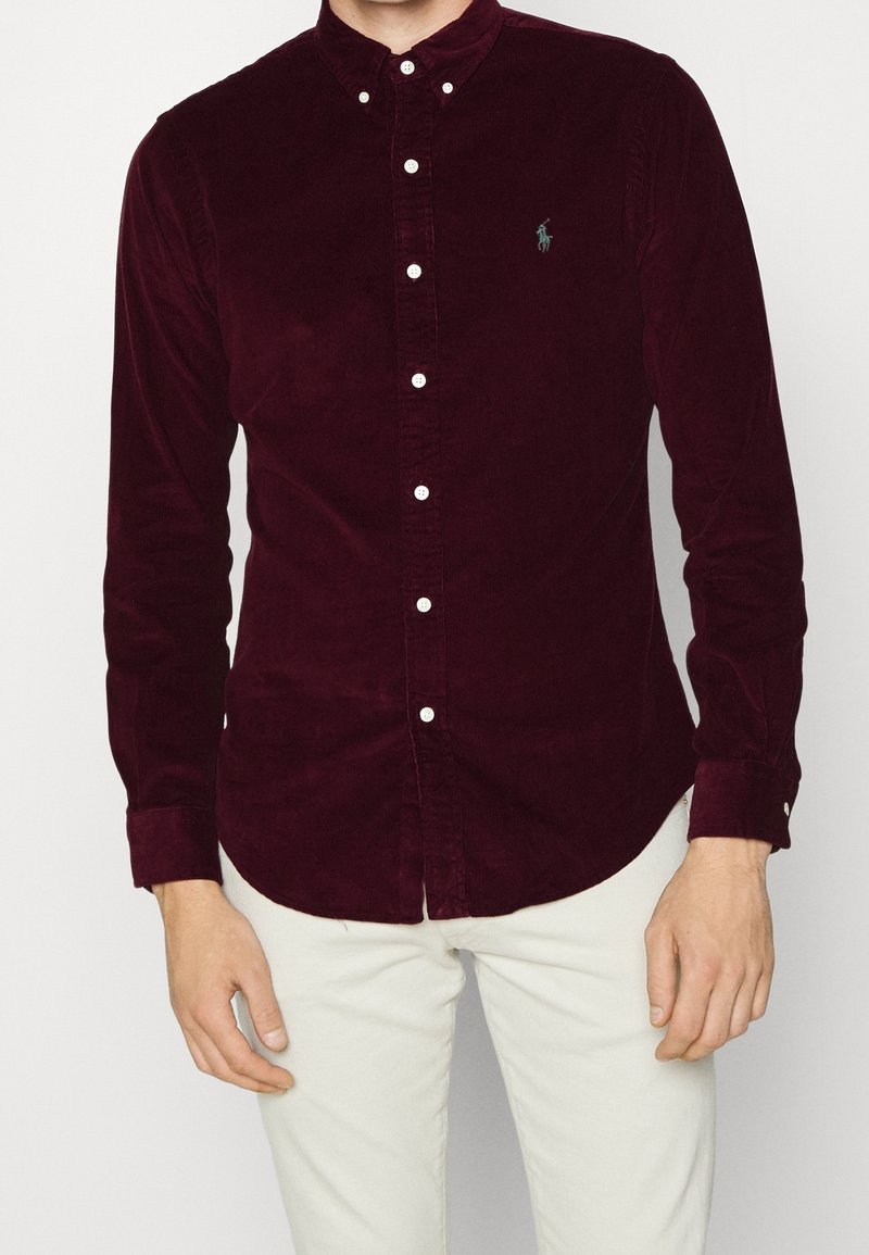 Hombre vistiendo una camisa de pana roja oscura con botones blancos y pantalones de colores claros, de pie contra un fondo liso.