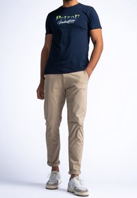 Tmavě modré bavlněné tričko s grafikou "PetroD Industries", doplněné světle béžovými úzkými chino kalhotami a šedobílými teniskami. Vyhrnuté nohavice kalhot.