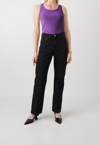 Kobieta w fioletowej, bezrękawnikowej bluzce, czarnych spodniach z wysokim stanem, czarnych szpilkach oraz złotych bransoletkach na prawej ręce.