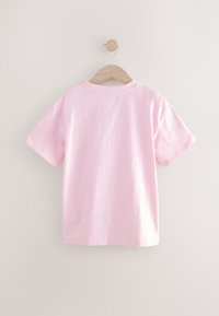 Roza bombažna majica s kratkimi rokavi, okroglim izrezom in sproščenim krojem, obešena na leseni obešalnik pred svetlim ozadjem.