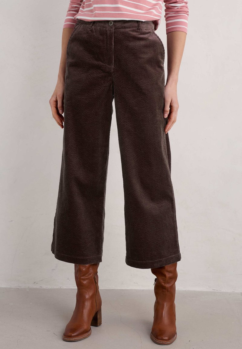 Hnědé kordové kalhoty širokého střihu s texturovaným povrchem, s knoflíkovým zapínáním a bočními kapsami. V kombinaci s hnědými botami na podpatku.