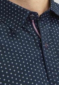 Camicia blu navy con un piccolo motivo a rombi bianchi, dotata di colletto a contrasto con dettagli a strisce rosse e bianche e bottoni neri.