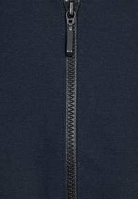 Marinblå zipjacka med en jämn tygtextur, centralkedja med svarta tänder och en blixtlåsdragning som matchar den övergripande färgen.