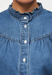 Gros plan d'une chemise en denim bleu avec un col à volants et des boutons blancs fixés le long de la patte de boutonnage avant.