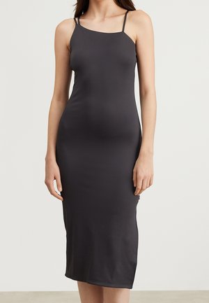 Vrouw draagt een aansluitende, mouwloze zwarte midi-jurk met dunne spaghettibandjes, staand tegen een effen lichte achtergrond.