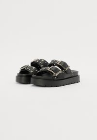 Sorte slidesandaler med to brede stropper prydet med sølvstifter og spenner, plassert på en kraftig, svart plattformsåle.