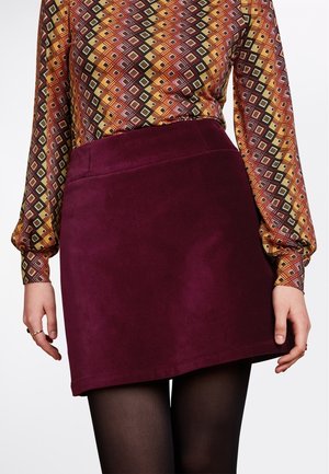 Femme portant un haut à manches longues à motifs rentré dans une minijupe bordeaux unie, collants noirs, mains posées le long du corps.
